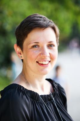 The image shows Laura Bavalics, a woman in a black shirt, with short brown hair, smiling against a green background