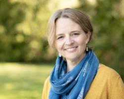 Headshot of woman with short blonde hair with yellow cardigan and blue scarf - grass and trees in background